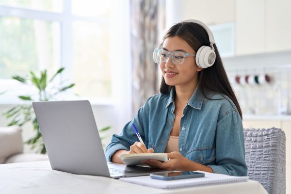 Woman wearing headphones and glasses taking notes while studying on laptop.