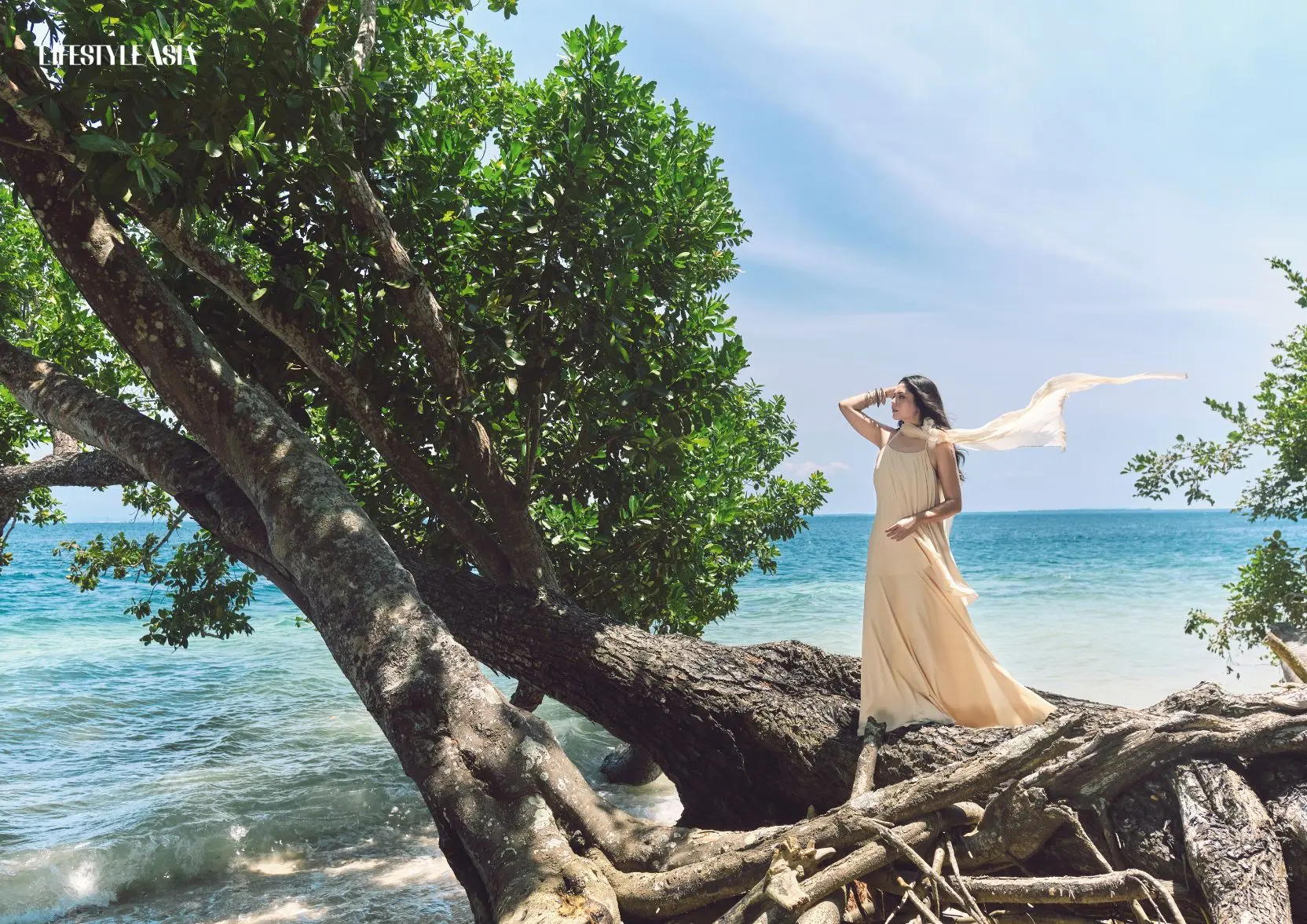 Woman in flowing dress standing on tree by the sea at tropical beach